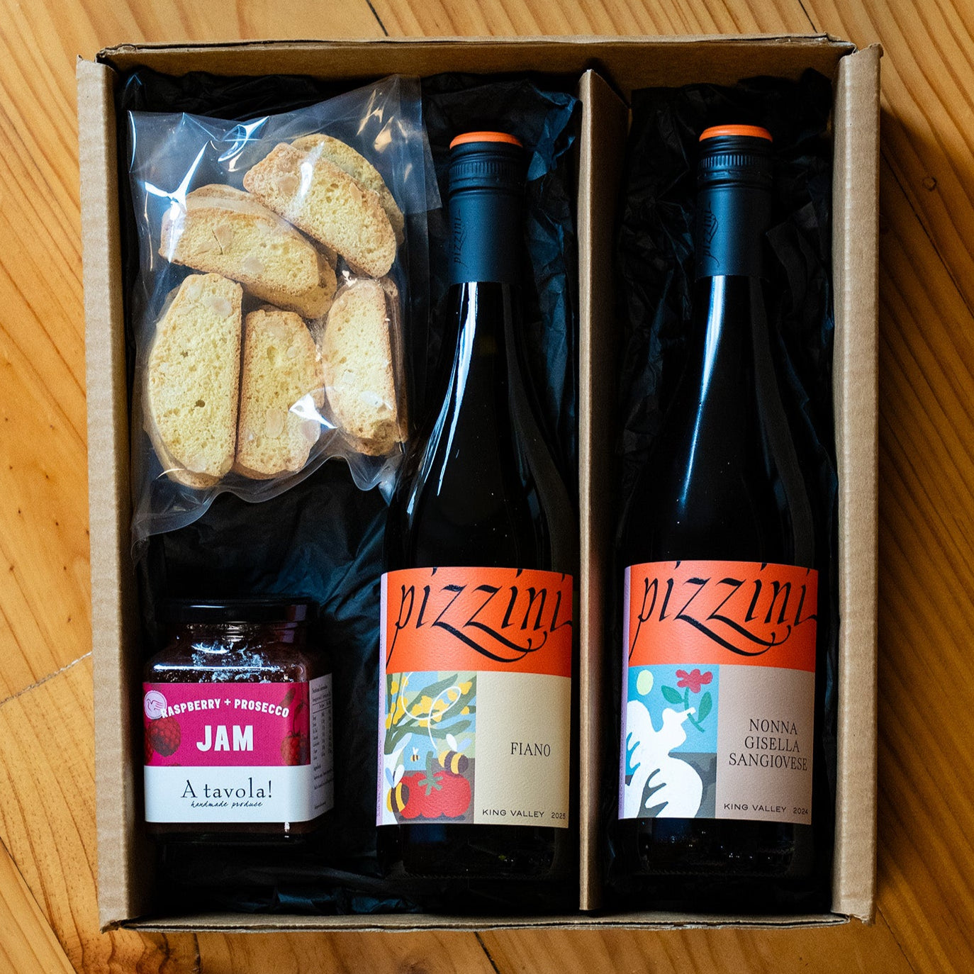 Gift set with two bottles of wine, a jar of jam, and some biscotti in a cardboard box on a wooden surface.